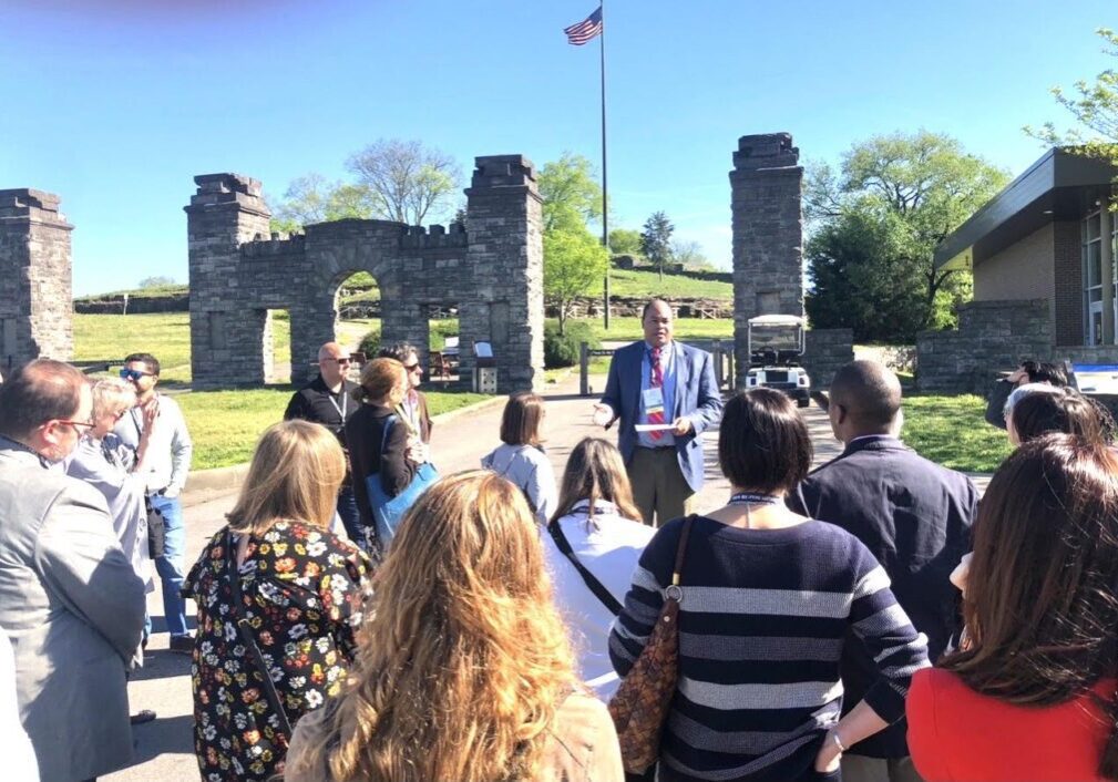A guide talks to a group near historic stone ruins on a sunny day.
