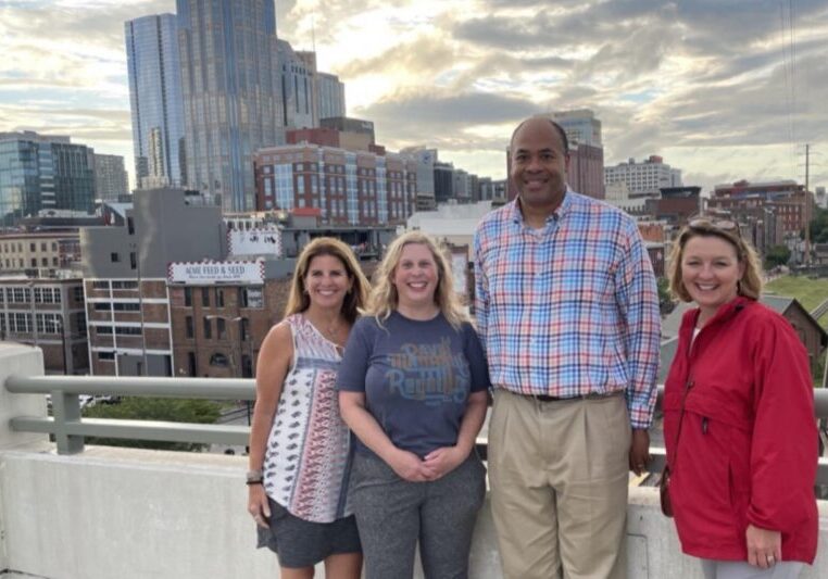 Five people posing on a rooftop with cityscape and cloudy sky behind.