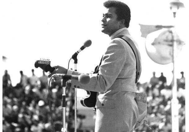 Black and white photo of a man playing guitar and singing on stage.