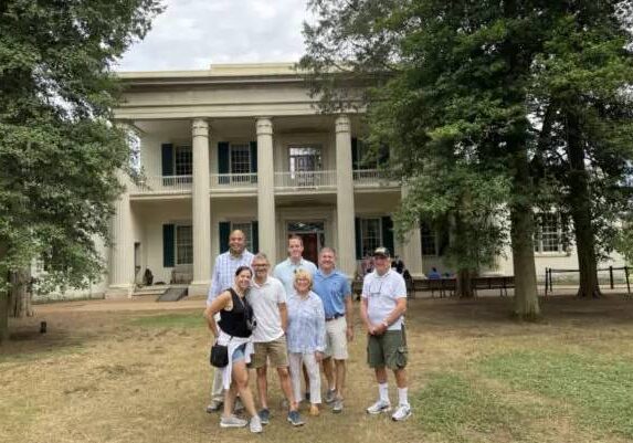A group of seven people posing in front of a large white building with columns.