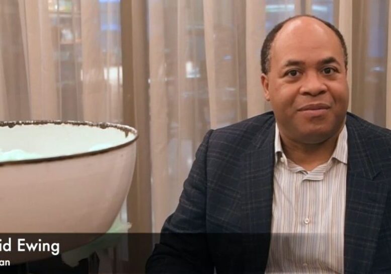 A man in a suit sitting beside a white chair and beige curtains.