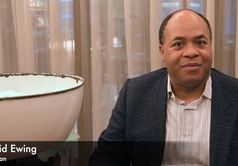 A man in a suit sitting beside a white chair and beige curtains.