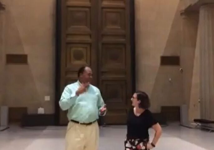 A man and a woman talking indoors near a large wooden door.