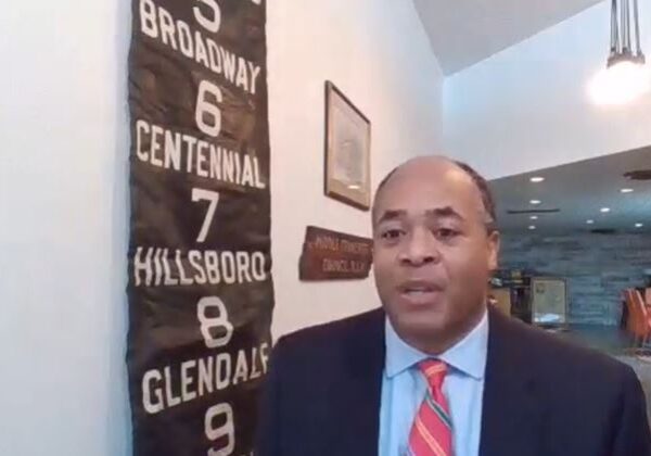 Man in suit and tie speaking indoors with a sports banner behind him.