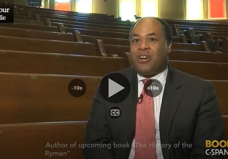 A man speaking in an empty auditorium with a video play button overlay.