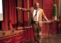 Man in a brown suit speaking and pointing on stage.
