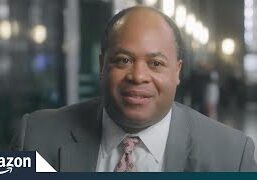 A man in a suit smiling indoors with blurred lights in the background.